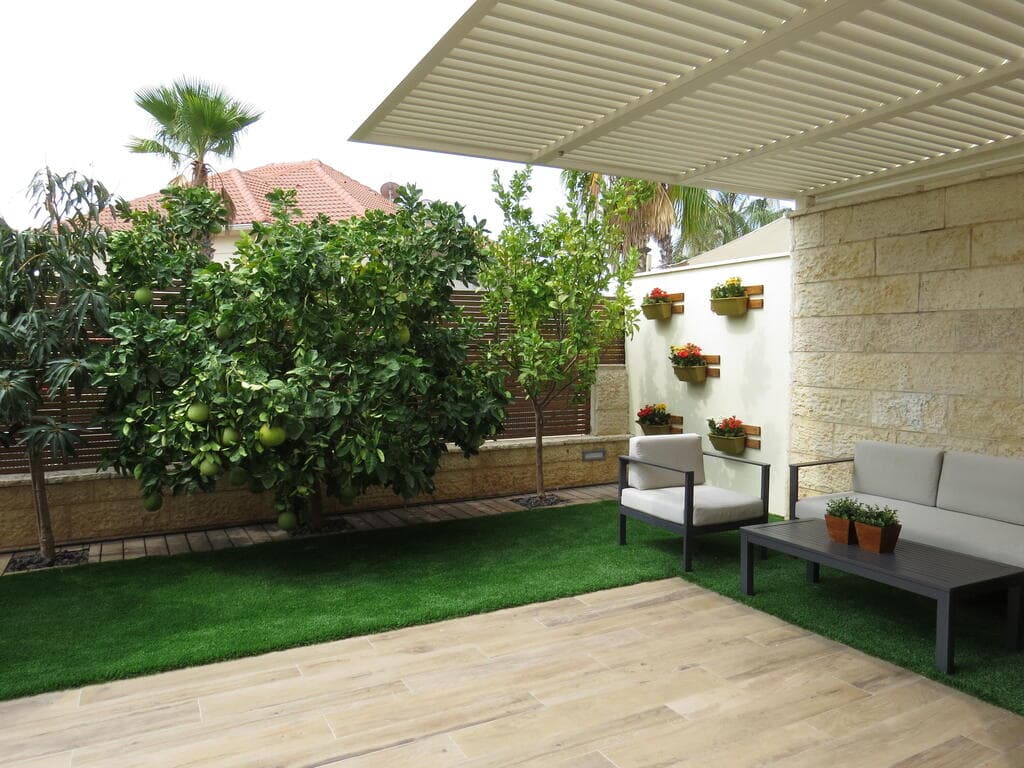 עיצוב גינה עם צמחייה מגוונת של עצי פרי וקיר אדניות שמשתלב עם הגדר (עיצוב וצילום: תמר זק) עיצוב גינה בדירת גן - צמחייה מגוונת. עצי פרי וקיר אדניות צמחים שמתכתב עם השפה העיצובית של הגדר