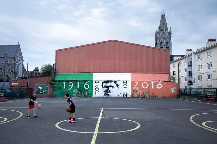 ילדים משחקים כדורסל כשמאחוריהם תזכורת לעימותים הפוליטיים שהובילו ליצירת המחתרת האירית
