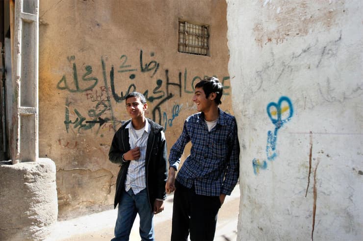 שני צעירים בשכונה יהודית בשיראז, איראן. על הקיר שמאחוריהם כתובת גרפיטי עם כינוי משפיל ליהודים