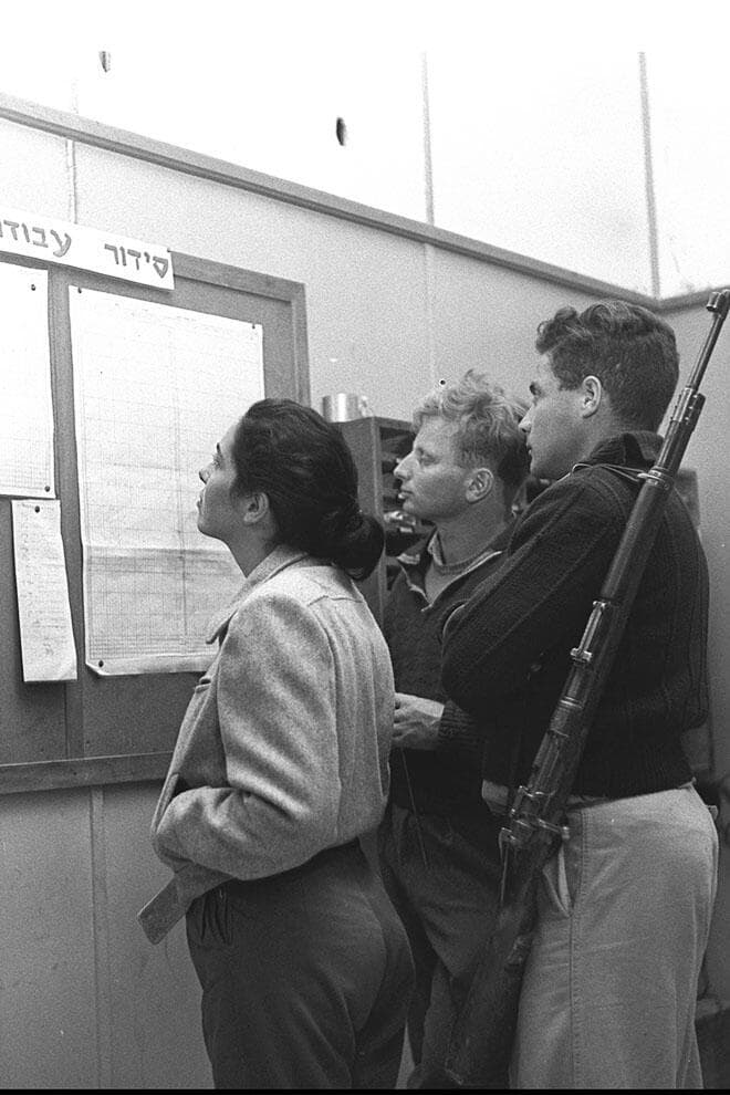 חברי קיבוץ נירים מול לוח סידור העבודה, בו יראו את שיבוצם לעבודה ביום או בשבוע הקרובים, 1955