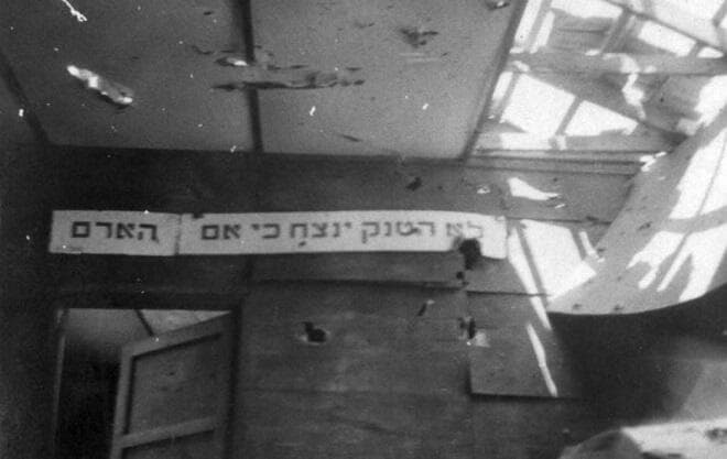 השלט על קיר צריף חדר האוכל היחידי שנותר עומד בסיום המתקפה, "לא הטנק ינצח כי אם האדם" מחגיגות ה-1 למאי 1948