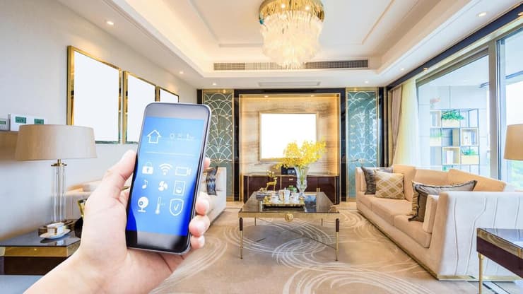 יותר ויותר מוצרים הופכים ל"חכמים" ונשלטים מרחוק (צילום: shutterstock) יד מחזיקה טלפון חכם על רקע סלון בית