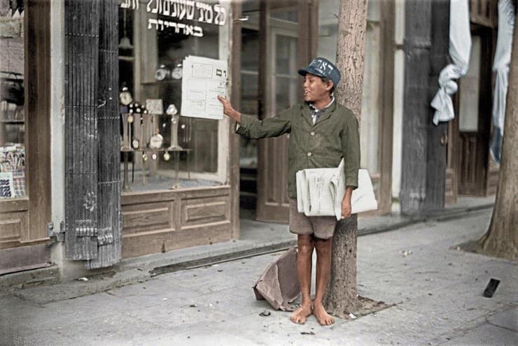 נער מוכר עיתונים ברחוב אלנבי, 1928 (צילום: יוסף שוויג, ארכיון הצילומים של קק"ל. צביעה: תמר הירדני) נער מוכר עיתונים ברחוב