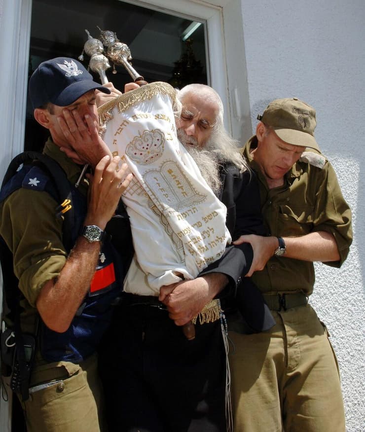 הרב פרומן מובל מחוץ לבית הכנסת ביישוב בדולח בגוש קטיף, בידו האחת אוחז בספר תורה ובידו השנייה מלטף את לחי החייל, 2005