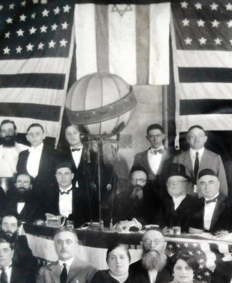 הרב קוק יושב במרכז, כשברקע דגל ישראל לצד דגלי ארצות הברית