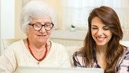 אישה מבוגרת יושבת ליד אישה צעירה ושתיהן מחייכות מול מחשב