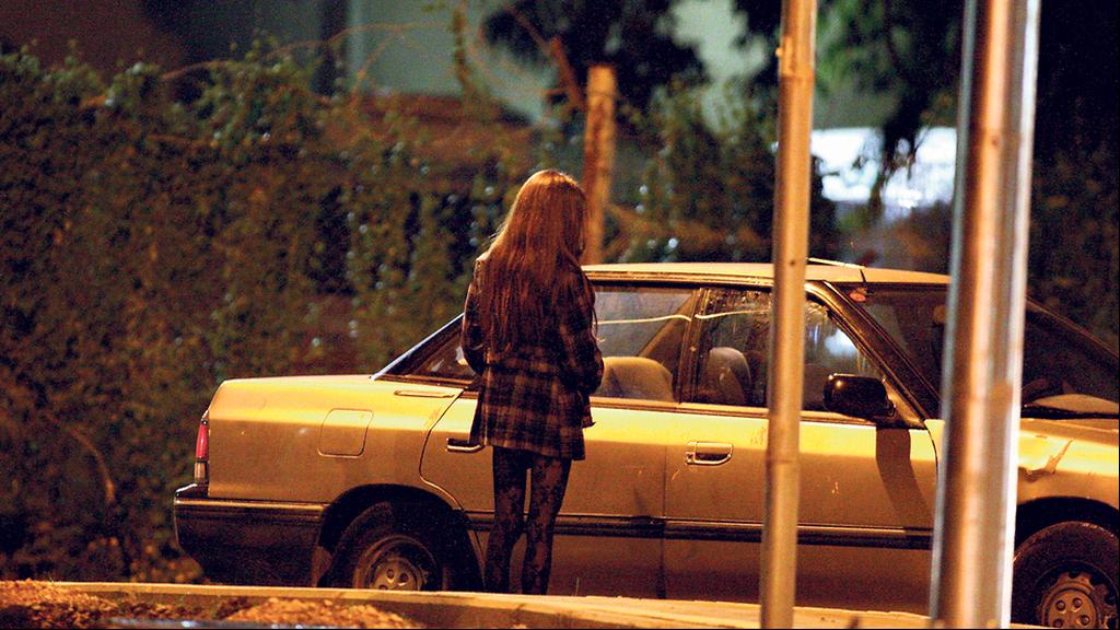 אישה בזנות עומדת עם הגב למצלמה ליד מכונית דוממת