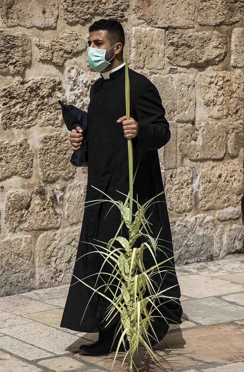 A monk wearing a face mask holds an olive branches outside the closed door of the Church of the Holy Sepulchre during the Catholic Palm Sunday 