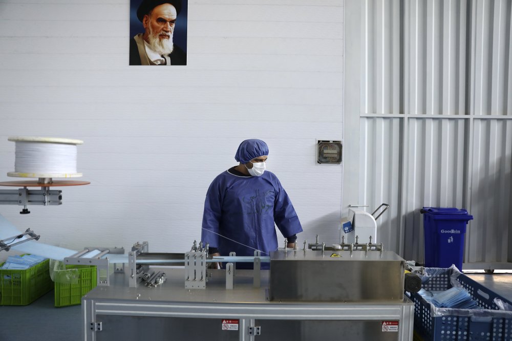 Under a portrait of the late Iranian revolutionary founder Ayatollah Khomeini, a worker wearing a protective suit controls a machine which makes face masks at a factory 