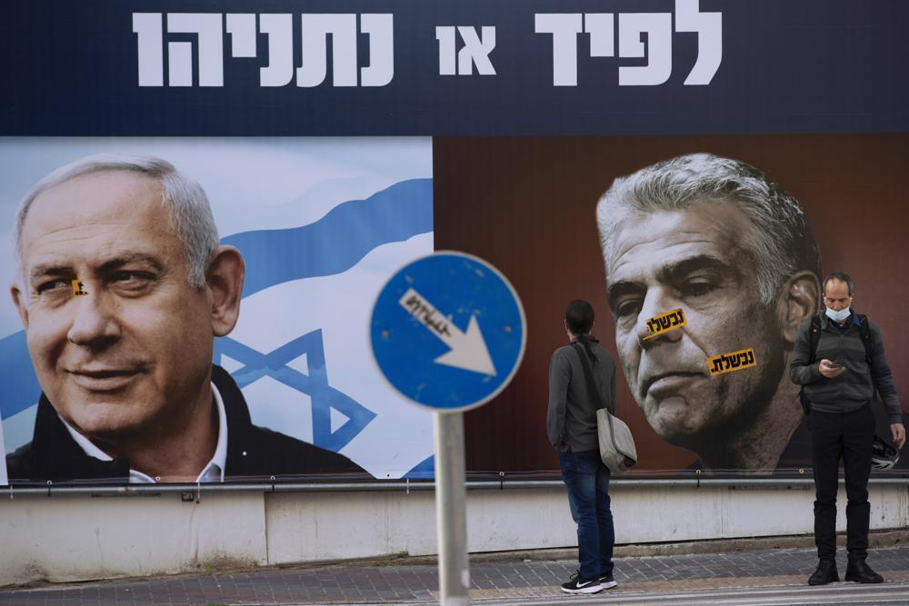 people stand in front of an election campaign billboard for the Likud party showing a portrait of its leader Prime Minister Benjamin Netanyahu, left, and opposition party leader Yair Lapid, in Ramat Gan, Israel 