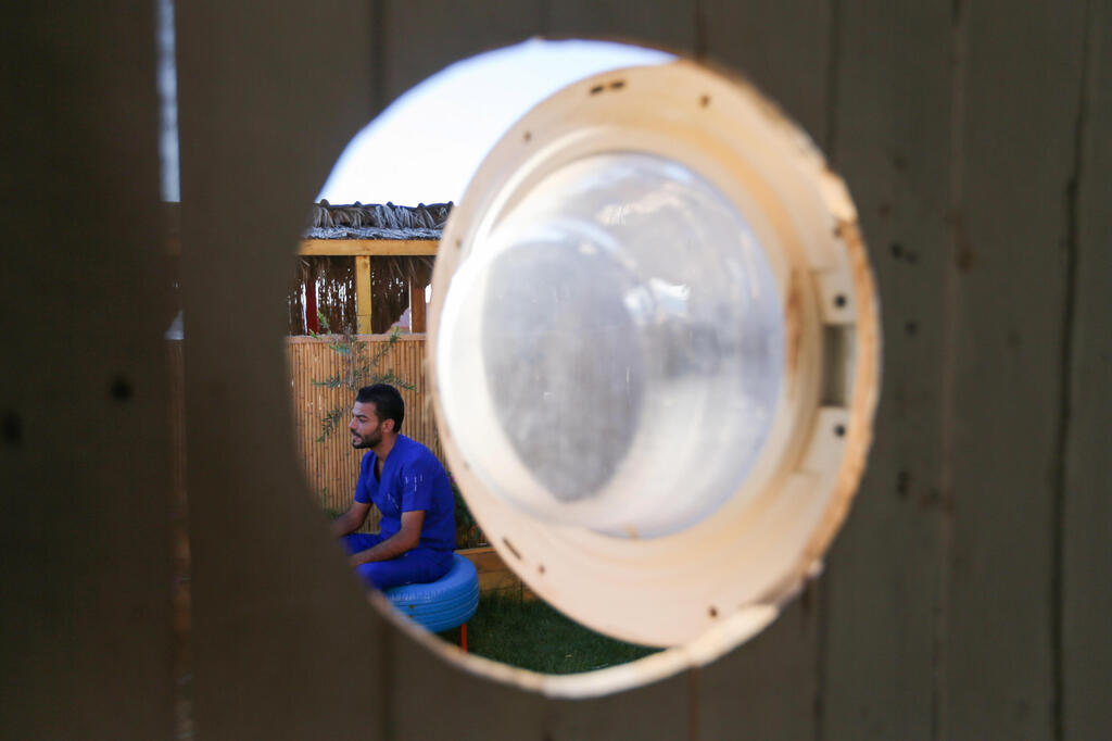 Palestinian artist, Ali Mhana, is seen through a glass door of a washing machine that he used as a window of a his makeshift office at an environment-friendly beachfront cafe in Gaza, July 8, 2021