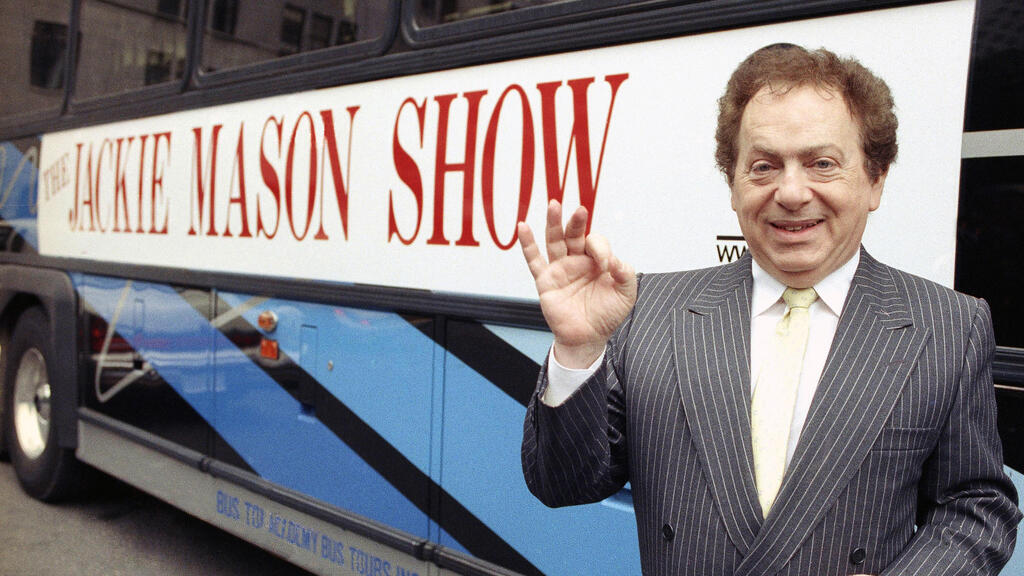 Actor/comedian Jackie Mason  stands beside a bus displaying a sign advertising his TV show,