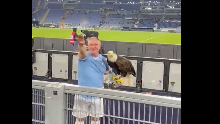  man holding a live eagle - Lazio's mascot- and wearing the blue and white colors of Lazio as he stands in front of the team's supporters at the stadium, raising his arm in the fascist salute