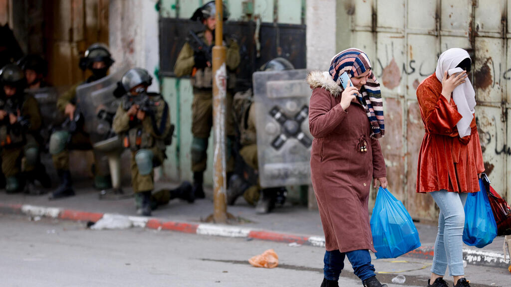 IDF forces during attest raid in West Bank as two Palestinian women walk by (Photo: Reuters) מחבל נהרג בעימותים בין פלסטינים לכוחות הביטחון צה"ל חברון