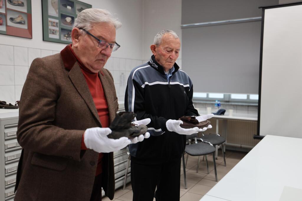 Auschwitz survivors Arie Pinsker (left center), and Bogdan Barnikowski, (right center) hold the shoes of child victims of the Nazis in the Conservation Lab in Auschwitz, joined by representatives of March of the Living, the Neishlos Foundation, and the Auschwitz-Birkenau Foundation.