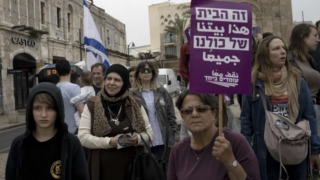 A Palestinian woman, center, takes part in a protest by Israelis in solidarity with Palestinians as part of ongoing protests against plans by Israeli Prime Minister Benjamin Netanyahu's government to overhaul the judicial system, in Jaffa, Israel, Thursday, March 23, 2023. The placard in Arabic and Hebrew reads: "This is home for all of us." 