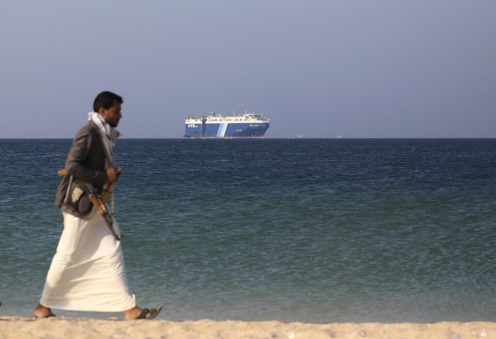 armed Houthi fighter walks through the beach with the Galaxy Leader cargo ship in the background, seized by the Houthis 