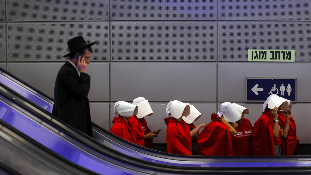 Protesters dressed as handmaids ride an escalator past an ultra-Orthodox man at Jerusalem’s central bus station during anti-government demonstrations in 2023 (Photo: Reuters) מחאה בירושלים