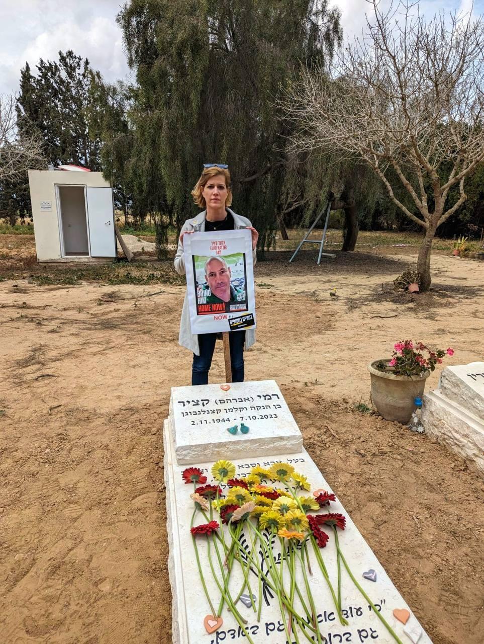 Carmit Palti Katzir holds a hostage poster of her brother Elad, who was killed in captivity, next to the grave of her father, who was murdered on October 7 כרמית פלטי קציר
