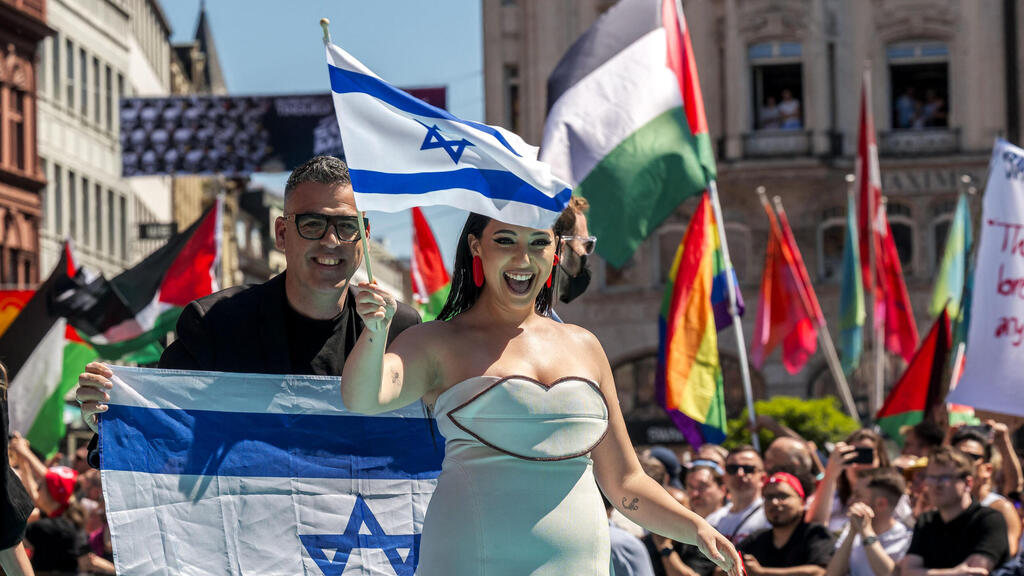 Yuval Raphael displaying Israeli flag as pro-Palestinian crowd protests behind her (Photo: Fabrice COFFRINI / AFP) יובל רפאל במצעד המשלחות של אירוויזיון 2025