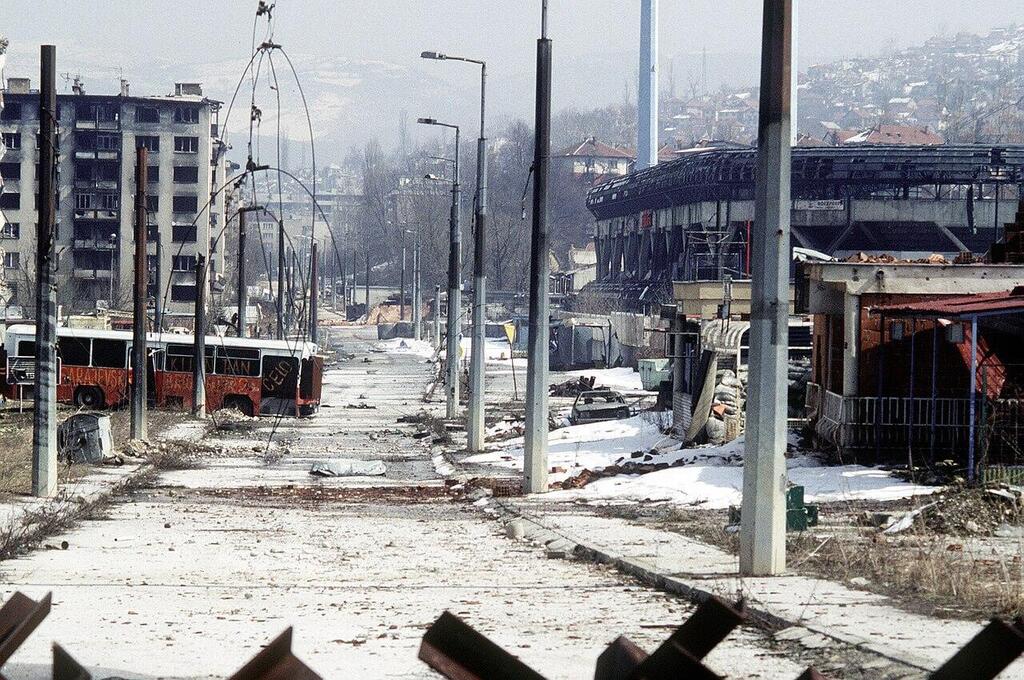 A street in Sarajevo lies in ruins during the Bosnian War, with damaged buildings and burnt-out buses visible in the background בוסניה סראייבו מצור מלחמה 1992