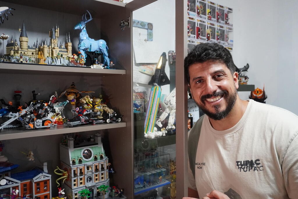 Stand-up comedian Matan Peretz poses next to his LEGO display at home, featuring detailed models from franchises like Harry Potter and Marvel 
