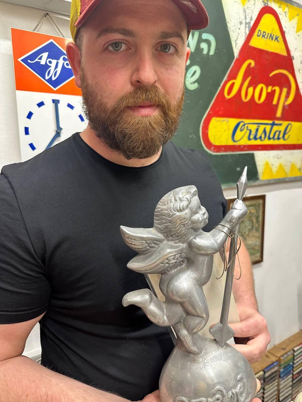Bar Gindy, host of the People & Collectors podcast, holds a vintage Cupid statue during a visit to collector Vital’s memorabilia archive. Behind him, a mid-century Cristal soda sign—one of the collection’s standout pieces—recalls the early days of Israeli consumer branding (Photo: Bar Gindy) Bar Gindy holds a vintage Cupid statue during a visit to collector Vital’s memorabilia archive. Behind him, a mid-century Cristal soda sign—one of the collection’s standout pieces—recalls the early days of Israeli consumer branding
