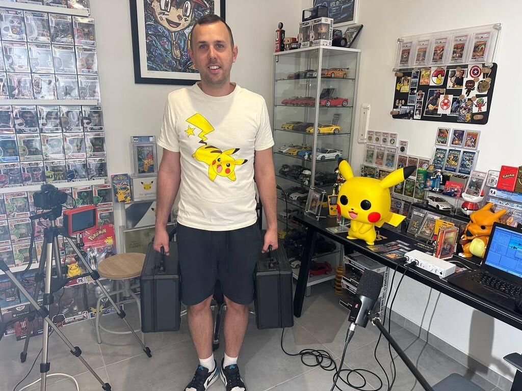 Dor, a longtime Pokémon collector and grader, stands in his collection room in Israel, holding two cases of trading cards. His space, filled with rare memorabilia, has become a hub for Pokémon collecting culture 