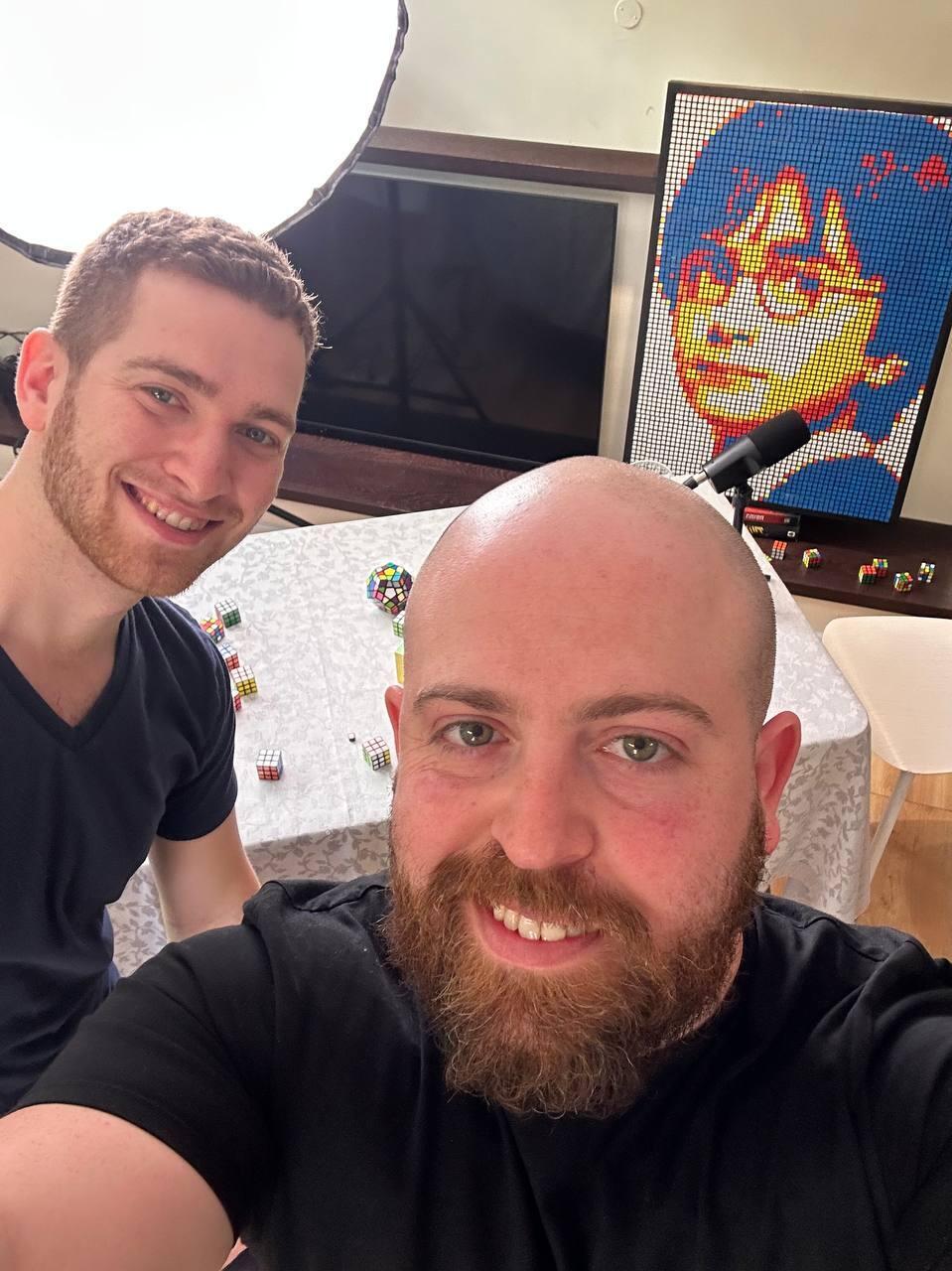 Tom, left, and Bar Gindy, host of the People & Collectors podcast, take a selfie in Tom’s home studio during a recording session. Behind them is a finished cube portrait built from hundreds of hand-arranged Rubik’s Cubes 