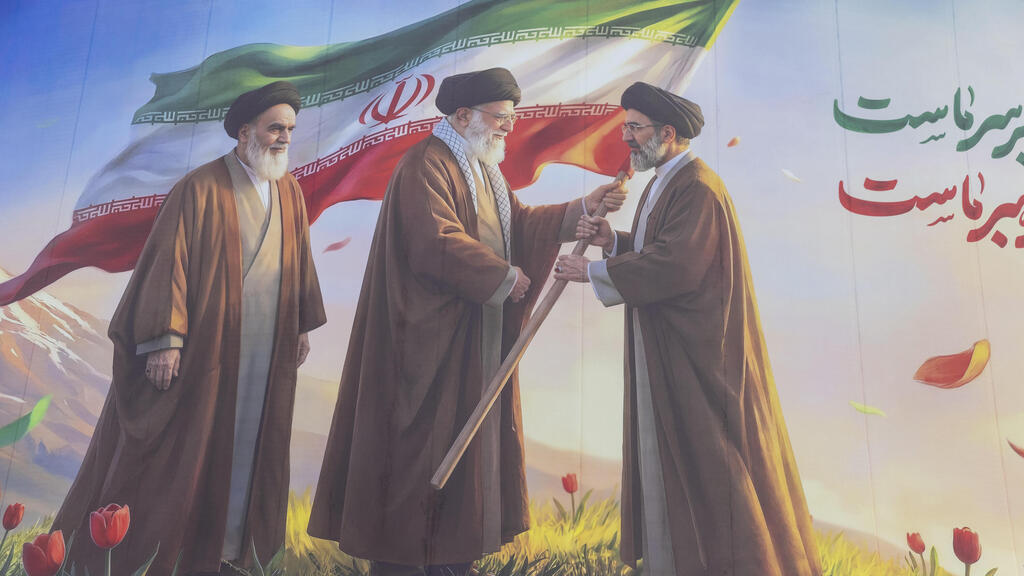 An Iranian poster depicting the transfer of the flag from Ali Khamenei to his son Mojtaba, alongside Ruhollah Khomeini (Photo: Majid Saeedi/ Getty Images) כרזה איראנית שמתארת את העברת הדגל בין עלי חמינאי לבנו מוג'תבא (לצד רוחאללה חומייני)