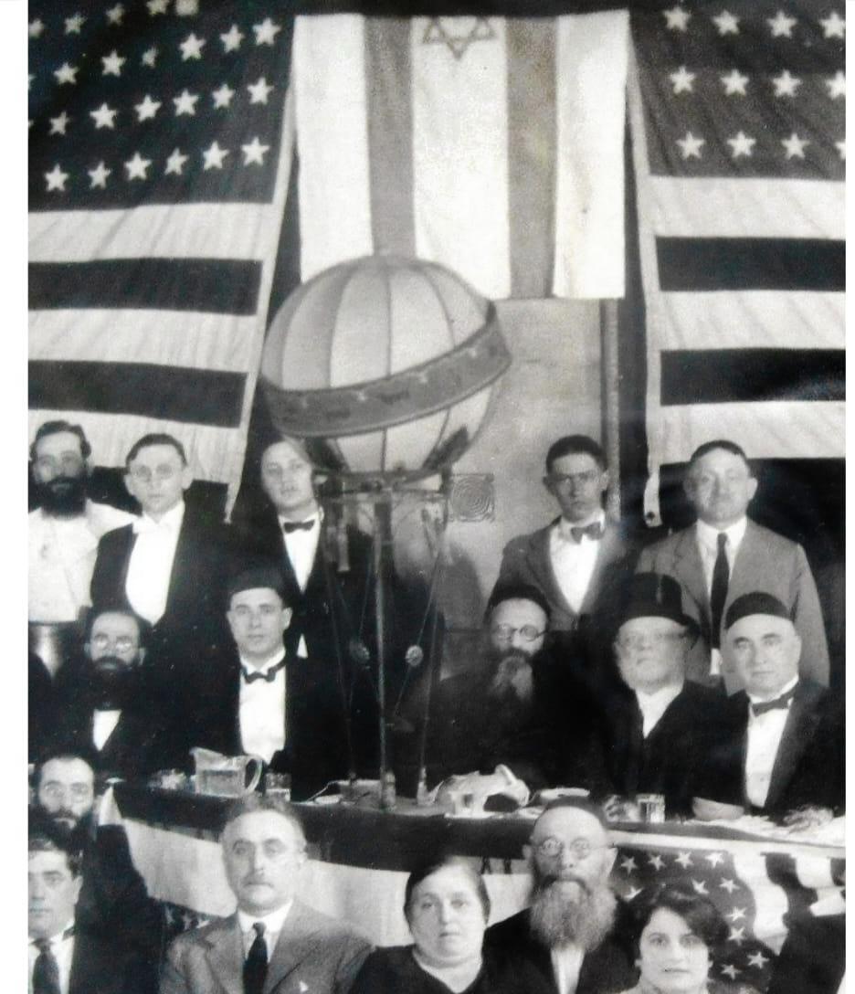 הרב קוק יושב במרכז, כשברקע דגל ישראל לצד דגלי ארצות הברית