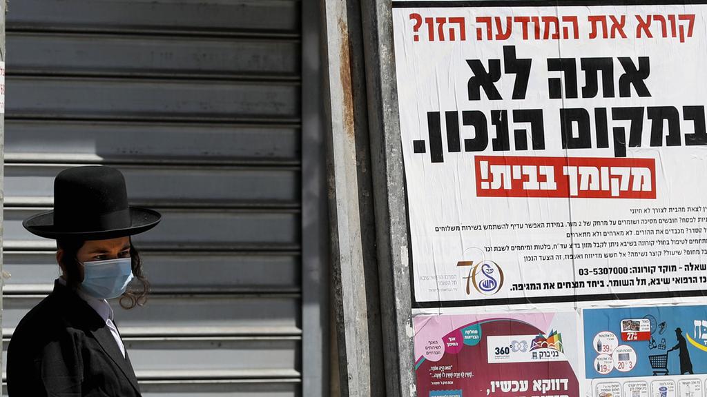 A Bnei Brak resident stands next to a poster urging residents to stay home. 'Reading this notice? You are in the wrong place,' the poster says
(Photo: AFP) A Bnei Brak resident stands next to a poster urging residents to stay home. 'Reading this notice? You are in the wrong place,' the poster says