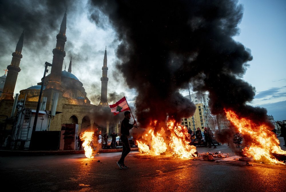 Protestors burn tires during a protest in downtown Beirut, Lebanon (Photo: AP) Mounir Hujairi, 23, holds up a Lebanese national flag as walks in front of burning tires set on fire to block a main road, during a protest in downtown Beirut, Lebanon