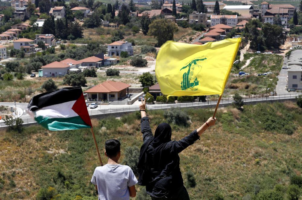 Lebanese wave Hezbollah and Palestinian flags, as they stand in front of the Israeli town of Metula, background, on the Lebanese side of the Lebanese-Israeli border in the southern village of Kfar Kila 