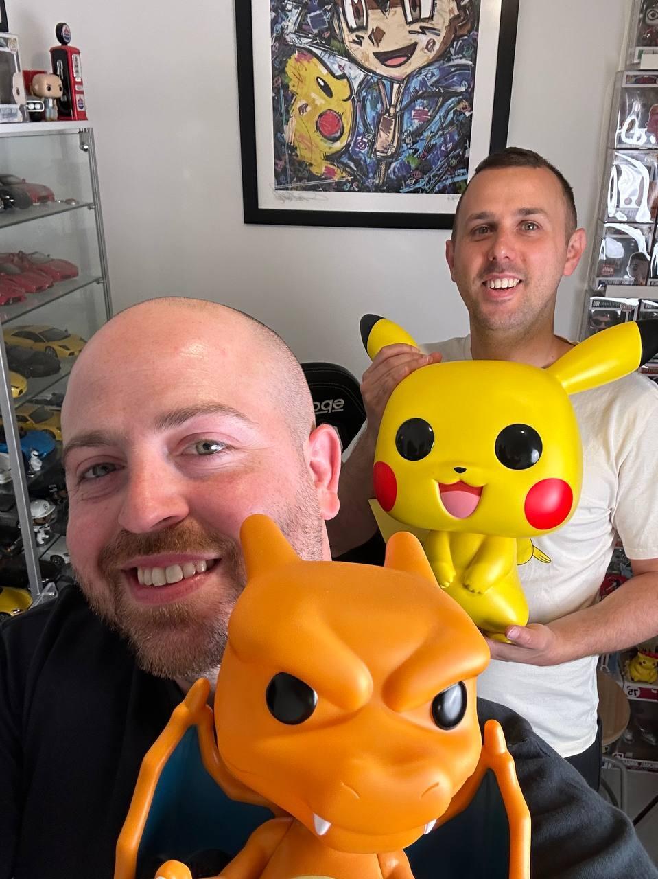 Bar Gindy, host of the People & Collectors Podcast, takes a selfie with collector Dor, holding oversized Pikachu and Charizard figures in Dor’s Pokémon collection room 