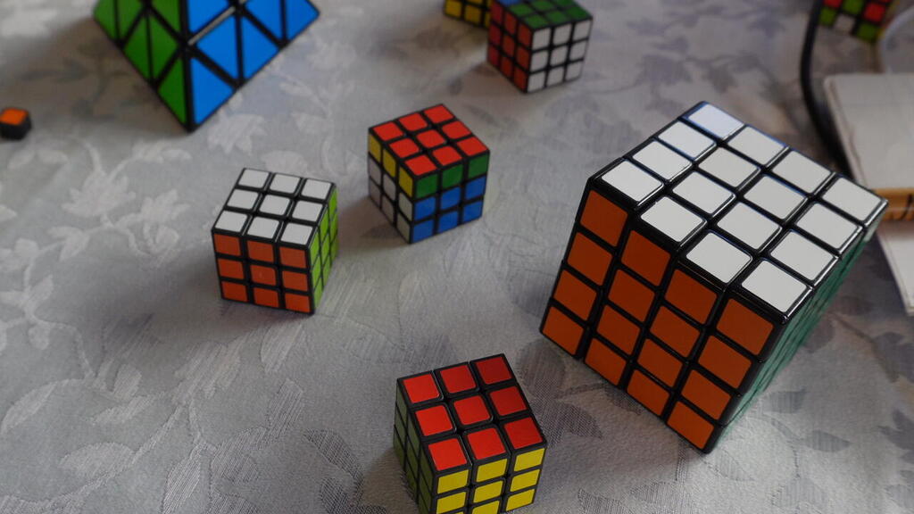 Various Rubik’s Cubes, including classic 3x3, 4x4 and pyramid variants, are scattered across a table at artist Tom’s home studio, where he creates large-scale portrait art by arranging and rotating each cube by hand to form pixelated images 