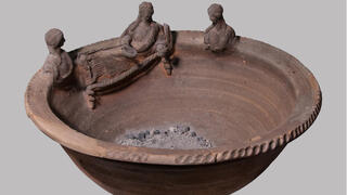 קטורת טרקוטה עם שאריות האפר שנחקרו. השפה מעוטרת בשלוש דמויות נשים, ככל הנראה מתארות אנשים שנפטרו לאחר מותם