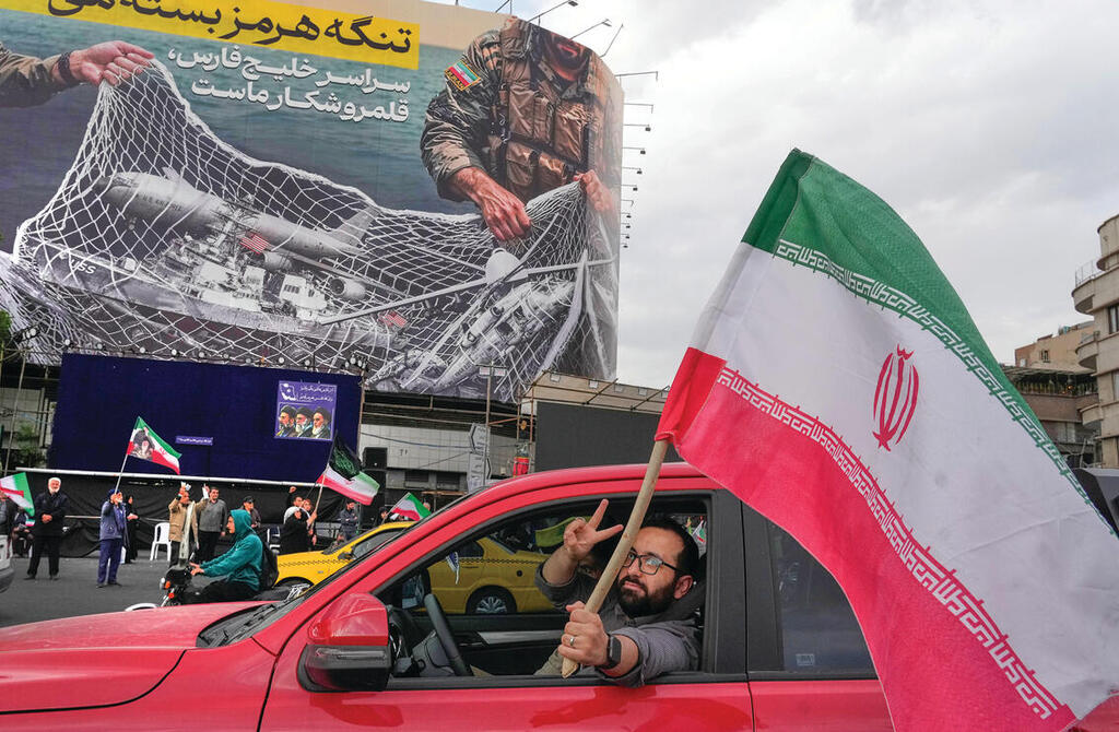 בתחושת ניצחון. אזרח איראני מסמן וי ומנופף בדגל מדינתו, מתחת לכרזת ענק בטהרן, שבה נראים מטוסים וספינות אמריקאיות נופלים ברשת האיראנית במצר הורמוז